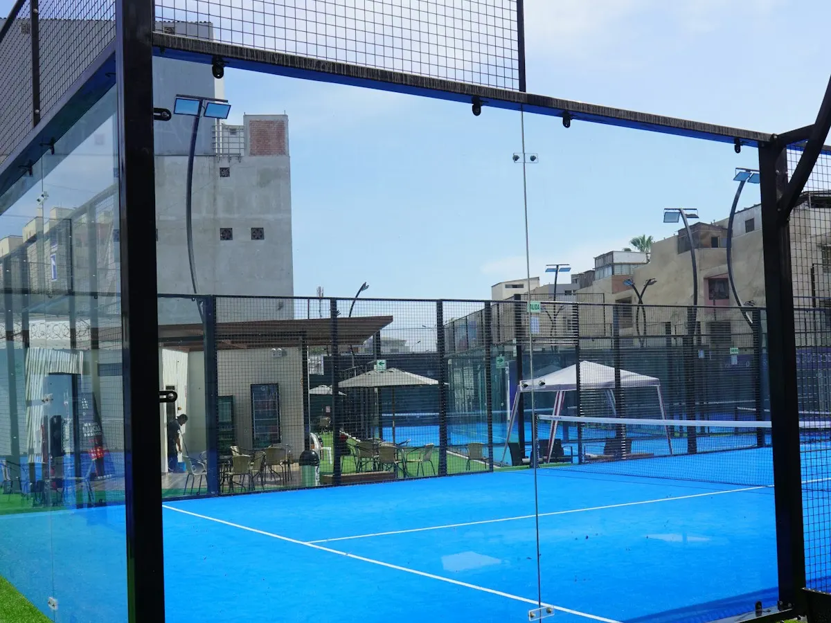 A padel court seen through its glass back wall, with metal mesh above and the net behind